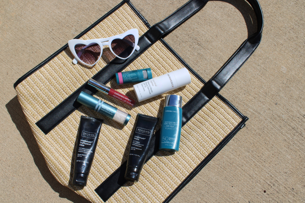 A straw tote bag displayed outdoors in natural sunlight, filled with a curated selection of mineral sunscreens including Revision Intellishade TruePhysical, Colorscience, and SkinMedica products, alongside white heart-shaped sunglasses — all recommended for post-laser skin protection.